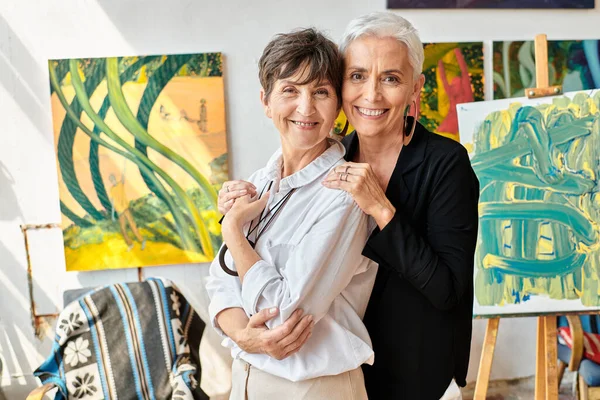 joyful mature lesbian artists embracing and smiling at camera near colorful paintings in workshop