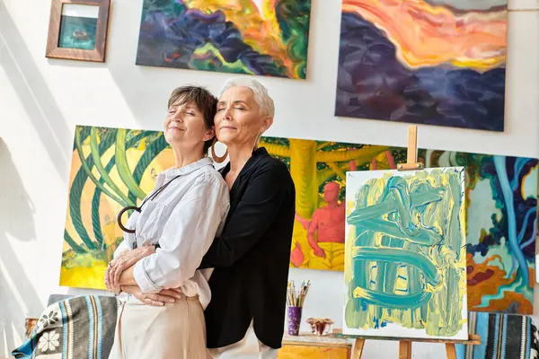 stylish and happy lesbian artists embracing with closed eyes near creative paintings in workshop