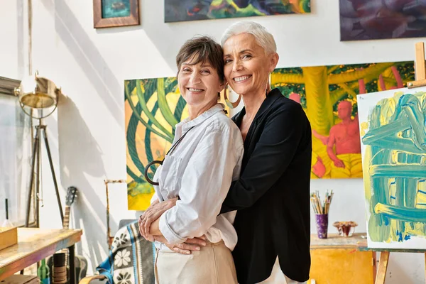 trendy and happy lesbian painters embracing and looking at camera near creative paintings in studio