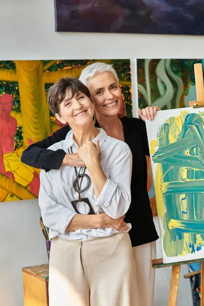 stylish and joyous lesbian painters embracing and looking at camera near paintings in workshop