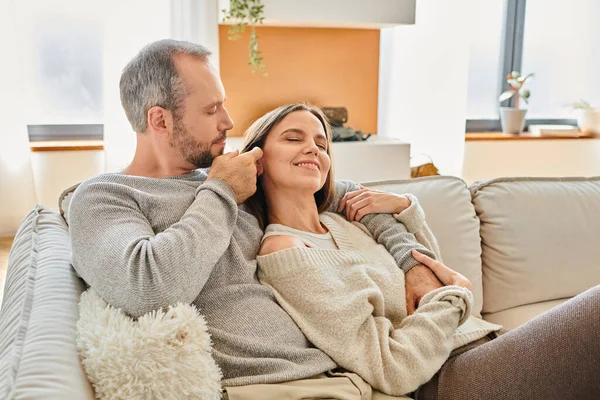 caring man fixing hair of pleased wife while sitting on cozy couch in living room, child-free couple