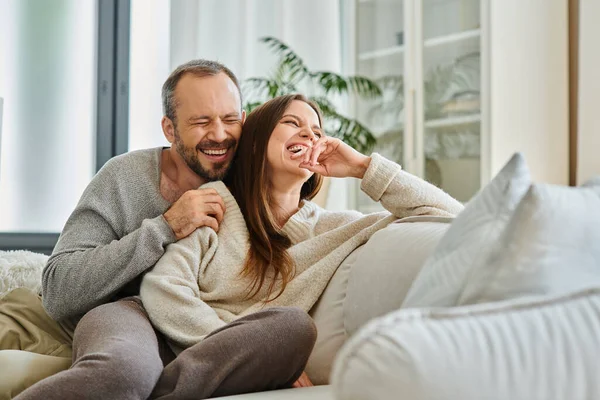 laughing man with closed eyes embracing wife on comfortable couch in living room, child-free couple