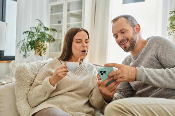 Man Visar Smartphone Till Förvånad Fru Med Kaffekopp Soffa Vardagsrummet — Stockfoto