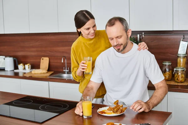 stock image caring wife holding orange juice near pleased husband enjoying breakfast in kitchen, love and care