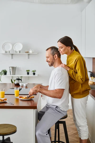 キッチンで妻の世話 子供なしのカップルの近くでおいしい朝食を楽しんでいる笑顔の男の側面の景色 — ストック写真