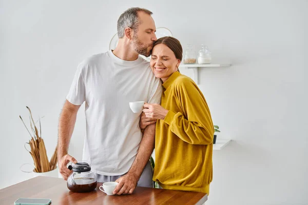 stock image husband kissing head of cheerful wife near fresh aromatic coffee in kitchen, child-free couple