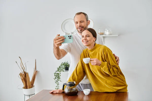 在现代厨房里 一对快乐的没有孩子的夫妇在早上喝咖啡的时候用智能手机自拍 — 图库照片