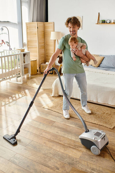 happy father multitasking housework and childcare, man vacuum uming bedroom with infant son in arms