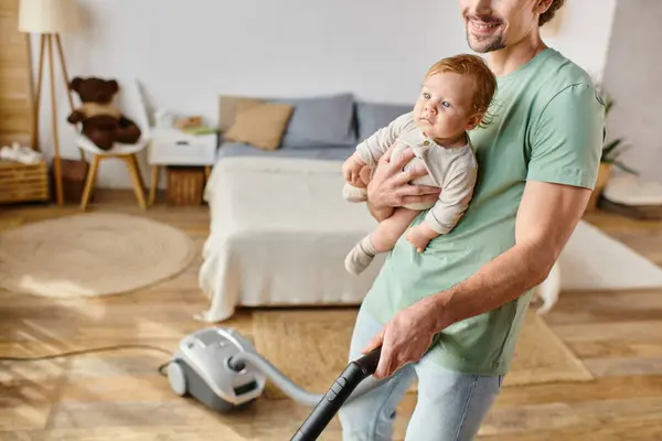 stock image cropped man multitasking housework and childcare, cheerful father vacuuming house with son in arms