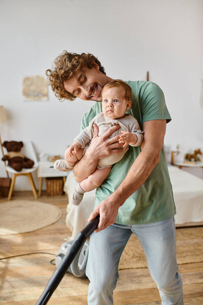 man multitasking housework and childcare, happy father vacuuming hardwood floor with baby in arms