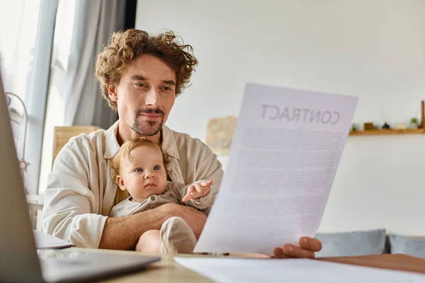 stock image busy man holding infant son and contract in hands while working from home, work-life balance