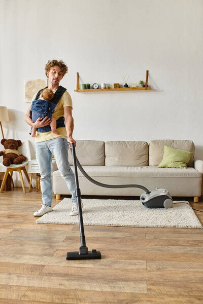 Curly father with infant baby boy in carrier vacuum uming hardwood floor living room, housework