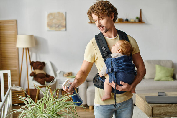 curly father with infant son in carrier holding spray bottle and watering green plant at home