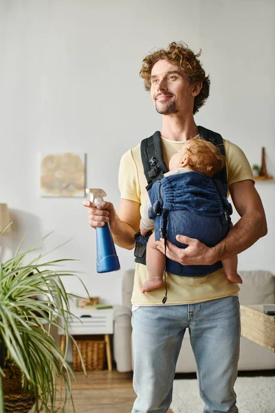 stock image happy father with infant son in carrier holding spray bottle and watering green plant at home