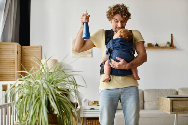 happy father looking at infant son in carrier holding spray bottle near green plant at home
