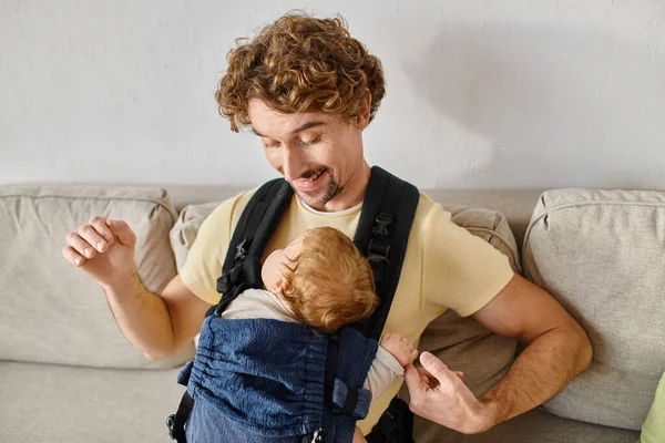 stock image cheerful man with curly hair looking at his infant son in baby carrier, fatherhood and love