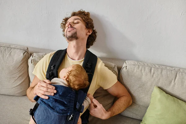 stock image curly haired father sleeping with infant son in baby carrier in living room, fatherhood and love