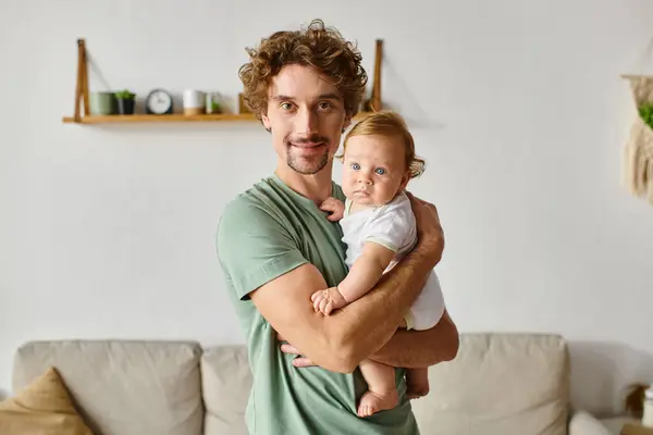 curly haired father with beard holding his infant son with blue eyes in a cozy living room, portrait