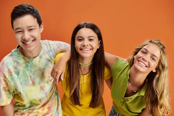 joyful multiracial teenagers in casual attires looking at camera on orange backdrop, friendship day
