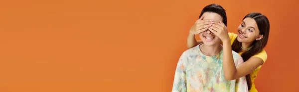 stock image pretty teenage girl closing eyes of her asian friend on orange backdrop, friendship day, banner