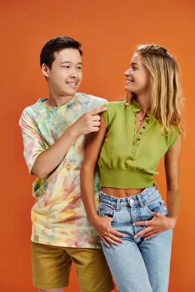 joyous interracial teenagers in casual attires smiling at each other on orange backdrop, friendship