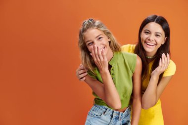 joyous adorable adolescent girls hugging and smiling at camera on orange backdrop, friendship day clipart