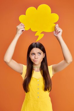 focused adolescent girl in everyday attire holding yellow thought bubble on orange background clipart