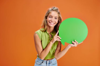 cheerful blonde teenager holding green speech bubble and looking at camera on orange backdrop clipart