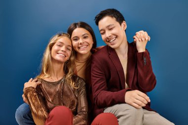 joyful teenage girls smiling at camera next to their jolly elegant asian friend on blue background clipart