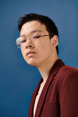 good looking asian teenage boy with glasses in stylish attire looking at camera on blue backdrop clipart