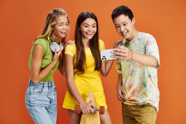 веселые юные друзья с наушниками и скейтбордом делают селфи на оранжевом фоне