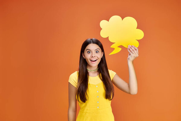 jolly teenager in vibrant attire holding thought bubble and looking at camera on orange backdrop