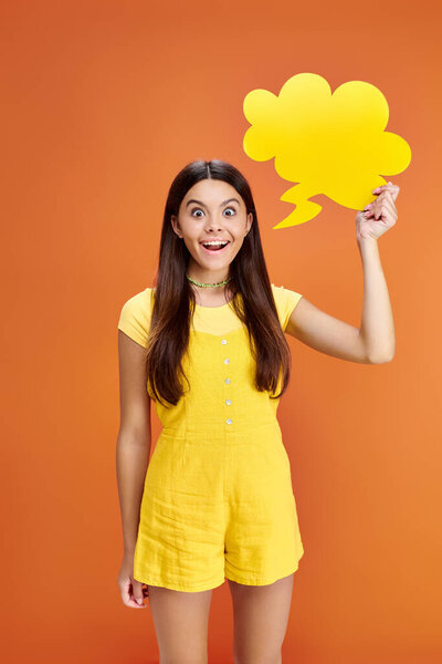 joyous teenage girl posing with thought bubble in hand and looking at camera on orange backdrop