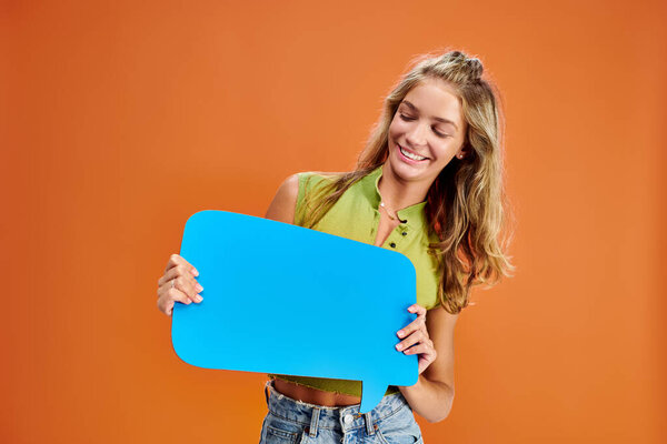 joyous blonde adolescent girl posing with blue thought bubble in her hands on orange backdrop
