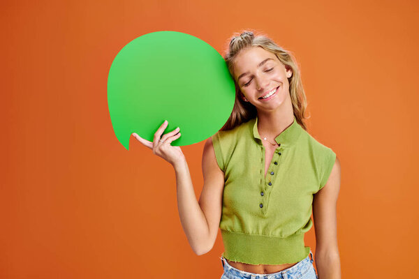 joyous teen in casual attire holding speech bubble and posing with closed eyes on orange backdrop