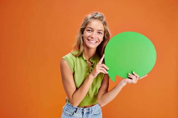 cheerful blonde teenager holding green speech bubble and looking at camera on orange backdrop