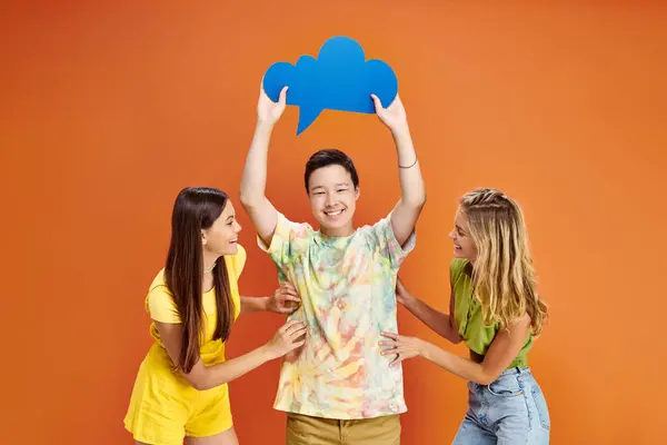 stock image three joyous diverse teenagers in vibrant attires posing with blue thought bubble on orange backdrop