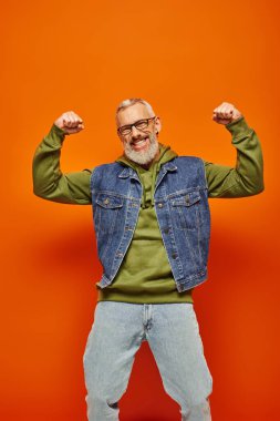 cheerful good looking mature man in denim vest posing with arms raised and smiling at camera clipart
