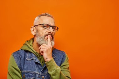 handsome focused mature man in green hoodie with glasses and gray beard posing on orange background clipart