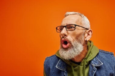 good looking mature man in green hoodie with glasses posing with open mouth on orange backdrop clipart