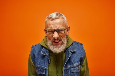 frustrated mature emotional man in vibrant green hoodie and denim vest posing on orange background clipart