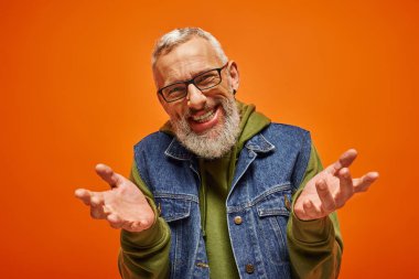joyous good looking man with beard and glasses shrugging and smiling at camera on orange backdrop clipart