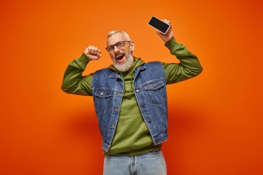 Yeşil kapüşonlu ve kot yelekli neşeli, çekici olgun bir adam elinde telefonla kameraya bakıyor.