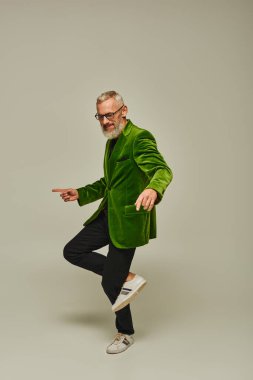 Yeşil renkli ceketli, olgun bir erkek model tek ayak üstünde mutlu bir şekilde gülümsüyor.