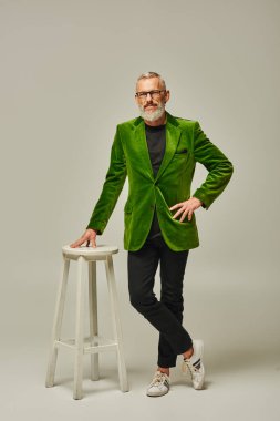 handsome mature funky man in green vivid outfit with glasses and beard posing near tall chair clipart