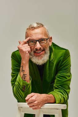 good looking joyous mature man in vivid attire posing with tall chair and smiling happily at camera clipart