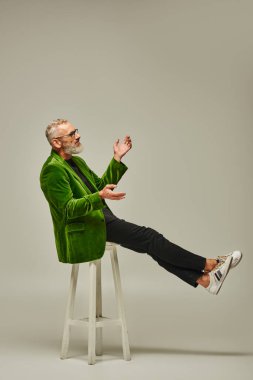 joyous mature man in green blazer with glasses sitting on tall chair in profile on beige backdrop clipart