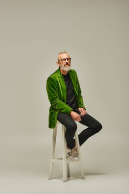 Sakallı, gözlüklü, uzun sandalyede oturan ve kameraya bakan olgun bir erkek model.