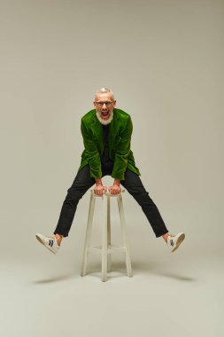 joyful good looking mature man in vibrant attire posing on tall chair actively and smiling at camera clipart