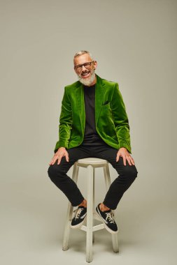cheerful bearded attractive mature man in green blazer sitting on tall chair and smiling at camera clipart